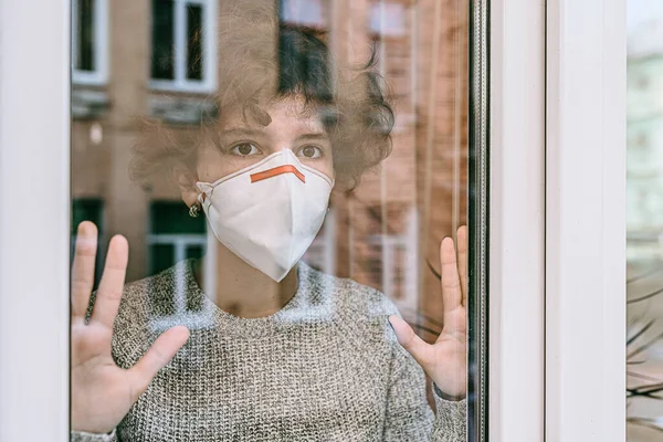 Kahverengi kıvırcık saçlı koruyucu maske takan genç kız pencereden dışarı bakıyor. Öğrenciler ve okul çocukları için karantina ve kendini izole etme. Camdan yansıyan ışık, bulanıklık, yansıma
