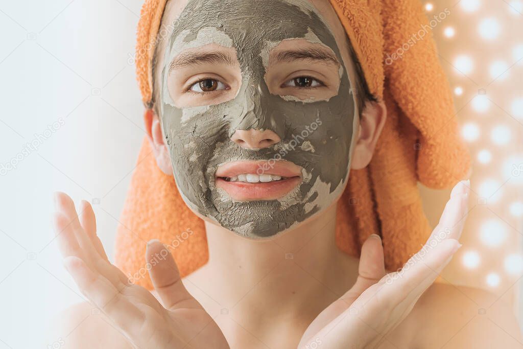Caucasian model girl teenager in towel on her head wearing cosmetic mask made of green healing clay on blurred background of lights of festive garlands, Teenager's facial skin care, spa salon at home