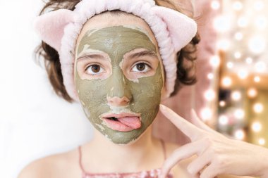 An attractive teenager girl in mask made of green clay makes faces on blurred background of Christmas garland. Spa treatments at home on eve of holiday. clipart
