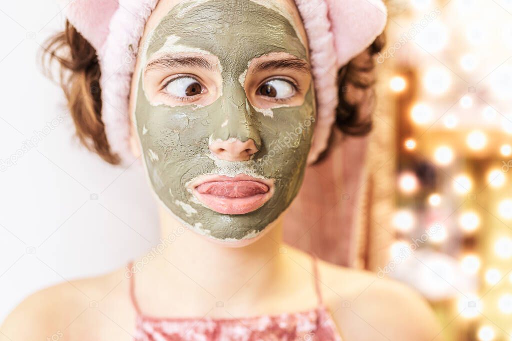 portrait of a teenage girl with brown hair with a gray clay cosmetic mask on her face with a funny grimace. Playful mood, make faces, slanted eyes. Beauty salon at home