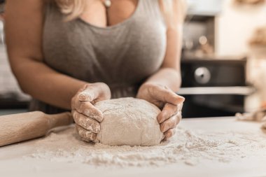 Artı, büyük göğüslü ve gri ev elbisesiyle derin dekolteli manken aile tatilinin arifesinde ya da Noel arifesinde ev mutfağında un kaplı ahşap kesim yüzeyinde elleriyle hamur yoğurur.