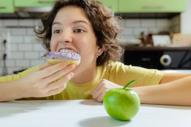 Teenage girl having fun eating donut. child chooses sweet unhealthy donut, ignoring green ripe apple. Teen girl with huge appetite eating bad donut