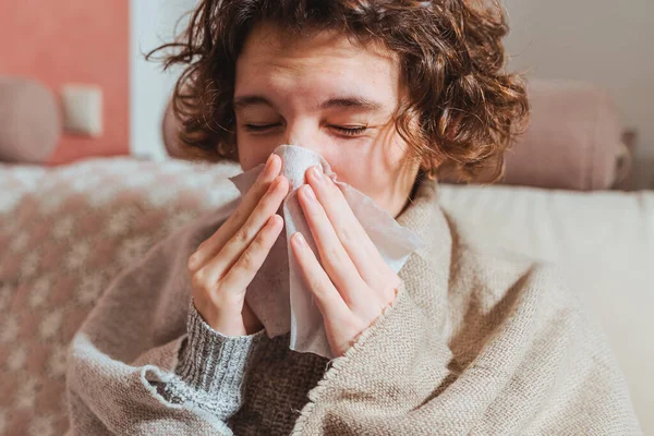 Sick upset young woman sitting on sofa covered with warm plaid, freezing, grimacing, runny nose, fever, sneezing into tissue, sick girl with flu symptoms, coughing at home, flu concept