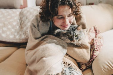 Yavaşça odaklan. Kahverengi kıvırcık saçlı, sıcacık battaniyeye sarılı genç bir kız tatlı, mırlayan Maine Rakun kedisine sarılıyor. Kedi yüzü ve genç kız yüzü. Kedileri ve insanları severim. İlişkiler, şefkat.