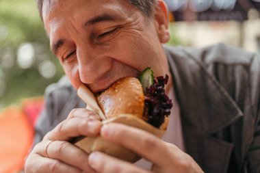 İştahlı orta yaşlı bir adam hamburger yer, zevkten gözlerini kapar. Fast food restoranında oturan adamın atıştırmalıkları var.