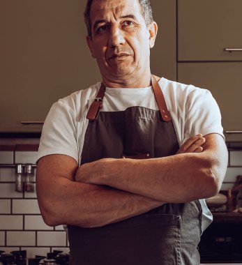 Orta yaşlı bir aşçı, önlüklü kaslı, göğsünde çapraz kollar olan, ev mutfağında duran. Erkek ev sahibi konsepti, ev mutfağının şefi. Kaslı adam mutfakta duruyor, hamur yapmak için malzemeler masada.