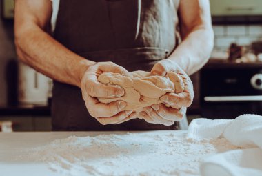 Erkek fırıncı, kahverengi önlüklü, hamur yoğuruyor güçlü kaslı kollarıyla, ev mutfağında duruyor. Ev yapımı hamurlar, ekmek, ev yapımı şehriyeler.