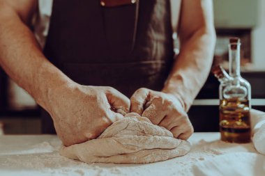 Erkek fırıncı, kahverengi önlüklü, hamur yoğuruyor güçlü kaslı kollarıyla, ev mutfağında duruyor. Ev yapımı hamurlar, ekmek, ev yapımı şehriyeler.