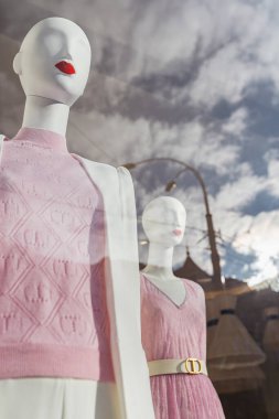 Parlak kırmızı dudaklı kadın mankeni, bahar koleksiyonu giysileri içinde, vitrinin arkasında, bulutların ve gökyüzünün yansımasıyla.