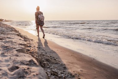 Sahilde gün batımında yürüyen tasasız mutlu bir kız. Genç, narin bir kadın deniz ya da okyanus kıyısında turist çantasıyla tek başına yürüyor ve gün batımını seyrediyor..