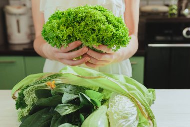 Genç kadın taze alınmış Lollo Bionda marulunu elinde tutuyor. Çevre çantasının içinde katlanmış yeşil sebze ürünleri mutfak masasında yatıyor. Mağazadan satın alınan faydalı vitamin sebzeleri. Kadın ev sahibesi, vitamin ve sağlıklı yiyecekler hazırlamak için eve getirdi.