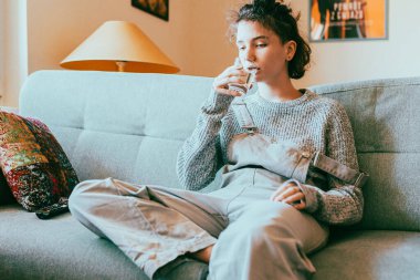 Kahverengi saçlı genç kız oturma odasında kanepede oturuyor ve bardaktan temiz su içiyor. Doğal kaynaklardan saf musluk suyu, filtrelenmiş su, susuzluk giderme. Sabahları temiz su iç, evdeyken ilaç al.