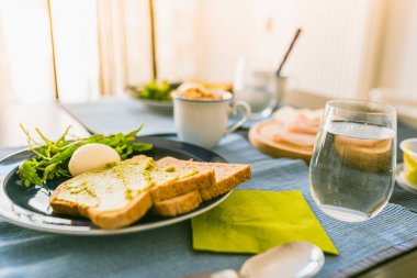 Guacamole soslu kızarmış ekmek, bir bardak temiz su, bir yumurta ve roka salatası. Kahvaltı için sağlıklı yemek. Bir apartmanın bulanık arka planında, masada pişmiş kahvaltı.