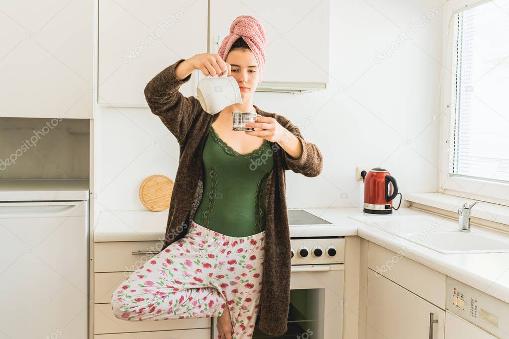 girl in home clothes balances on one leg, standing in home kitchen, with bath towel on her wet hair, pours coffee into cup. Morning coffee in yoga pose at home of teenage girl