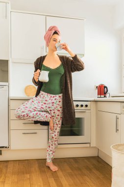 Evde giysi giyen kız bir bacağının üzerinde duruyor, mutfakta ıslak saçlarında havluyla duruyor, fincandan kahve içiyor, diğer elinde süt testisi tutuyor. Yoga pozunda sabah kahvesi. Evde genç bir kız pozu.