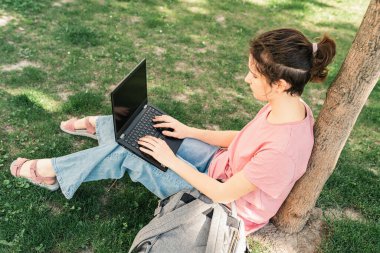 Genç bir kız, spor salonu öğrencisi ya da öğrencisi, yeşil çimlerde dizüstü bilgisayarla oturuyor, ödev yapıyor, makale yazıyor, online ödevlerini tamamlıyor. Gençlerin modern teknoloji ve aletlerinin kullanımı