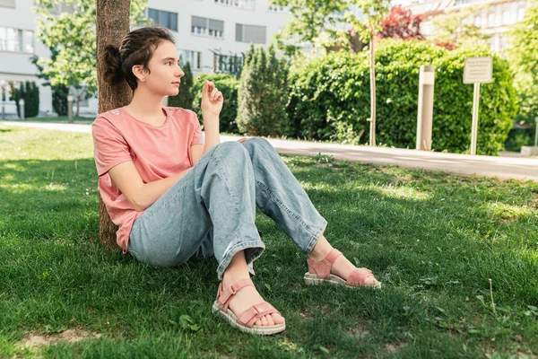 attractive teenage girl sits in park on lawn, holds small chamomile flower in hands, thoughtful emotion on face, thoughts of happiness. Positive emotions, mental and emotional health