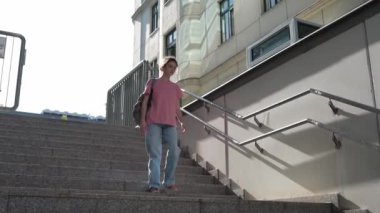 Sırt çantalı genç bir kız, günlük giysiler içinde güneşli bir günde merdivenlerden aşağı iner, aceleyle üniversiteye gider, merdivenlerden yukarı çıkar.