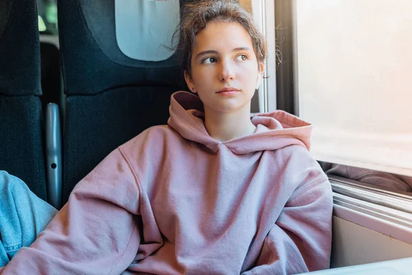 beautiful girl, in sweatshirt, with hair in bun, with dreamy expression, rides train, enjoys  trip alone, looks out window. Enjoy an exciting ride alone. Student on vacation travels around Europe