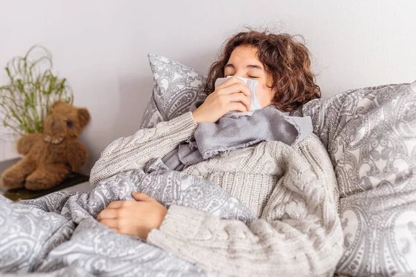 Colds, sneezing, viral infection, allergic reaction to pets, dust, or houseplants. Teenage lying in bed, with warm scarf wrapped around throat, in knitted sweater, sneezes covering mouth with napkin