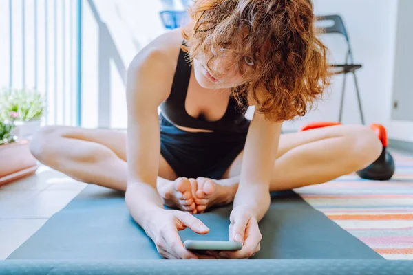 young lady teenager in sports top shorts sits on gymnastic mat, butterfly pose, stretches muscles before online workout, at home on open loggia. In hands of smartphone. Butterfly pose muscle training