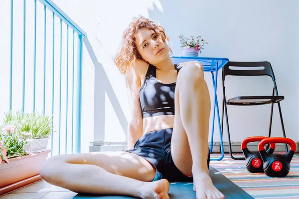 teenage girl in sports top and shorts sits on yoga mat, on loggia in sun, resting after workout. Young sports girl after sports workout home, without leaving home, sports equipment weights for pumping up muscles