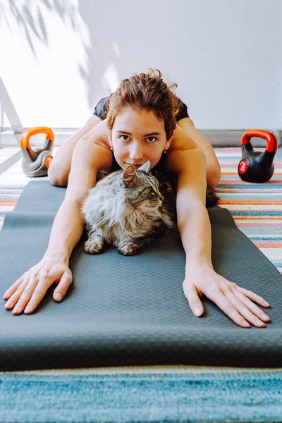 Home stretching workouts with pets. sporty teenager sitting on mat in kundalini pose, stretching back muscles, legs in butterfly pose, doing breathing exercises, next to beloved cat, mental health concept