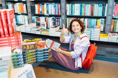 Üniversite öğrenci kütüphanesindeki genç öğrenci eğitim edebiyatının yanında oturuyor, başparmağıyla olumlu işaret veriyor. Z jenerasyonu kağıt kitapları e-kitaplara tercih eder. Viyana, Avusturya 06 2022