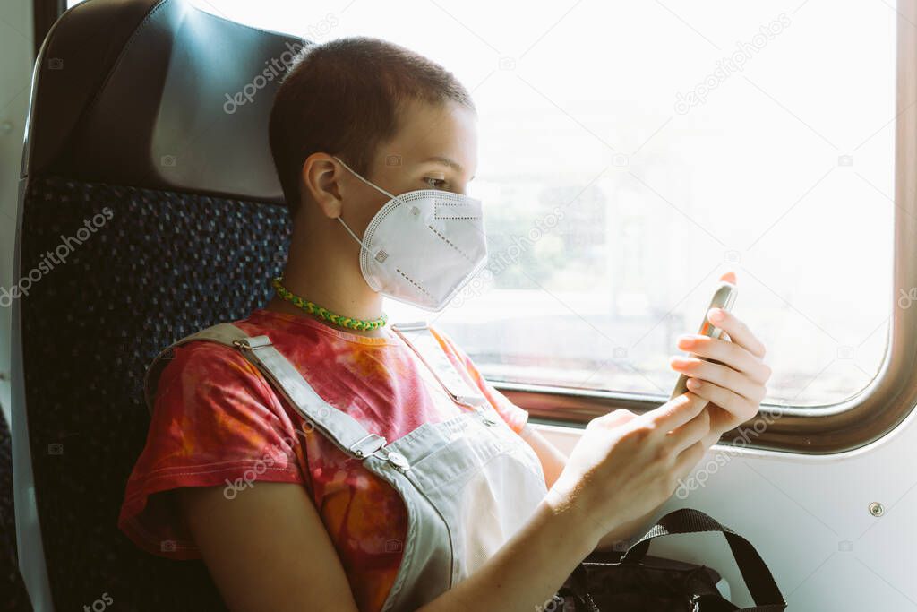 New norms for train travel in Europe. beautiful thin young girl with shaved hair, in protective mask, travels alone by train, uses smartphone to communicate, sitting at window of car
