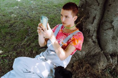 Genç kız ağacın altında çimenlikte oturur, gövdeye yaslanır, internetten yemek pişirir, görüntülü sohbet izler, selfie7 çeker.