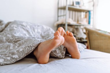 legs of young woman peek out from under covers. lying in bed under warm blanket in hot weather, sound sleep, children's healthy sleep, circadian rhythms concept. Lovely legs peek out from under covers on bed. Side view.