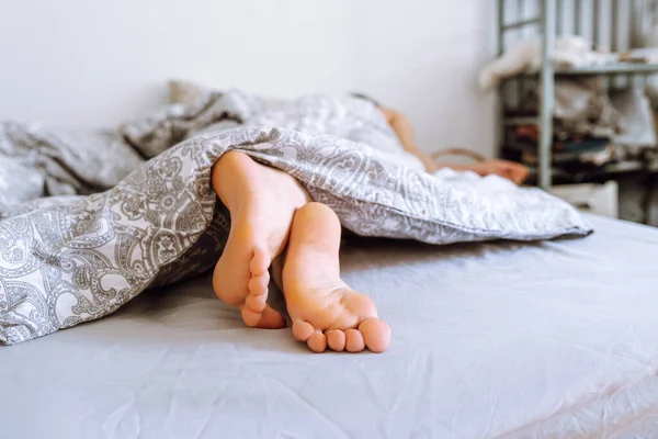 legs of young woman peek out from under covers. lying in bed under warm blanket in hot weather, sound sleep, children's healthy sleep, circadian rhythms concept. Lovely legs peek out from under covers on bed. Side view.