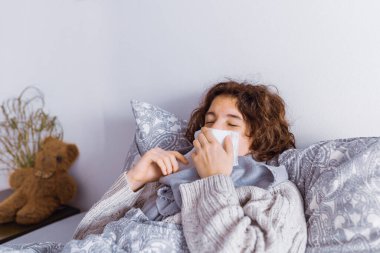 Yatakta uzanan genç, boğazında sıcak bir eşarp örgü süveter, ağzında peçeteyle hapşırıyor. Soğuk algınlığı, hapşırma, viral enfeksiyon, evcil hayvan, toz ve bitkilere alerjik reaksiyon