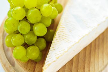 Brie peyniri, tahta kesme tahtasındaki beyaz üzümler. Yan görüş, yakın çekim. Brie peyniri ve bir demet beyaz üzüm.