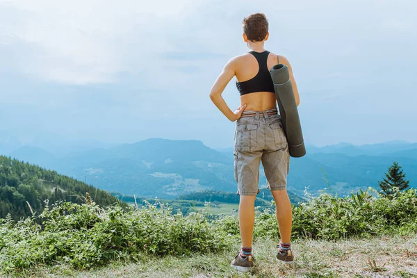 teenage girl athletic build, with short haircut, in sports top and shorts, with yoga mat, stands on edge cliff and admires mountain landscape, view from back. woman hiking in mountains
