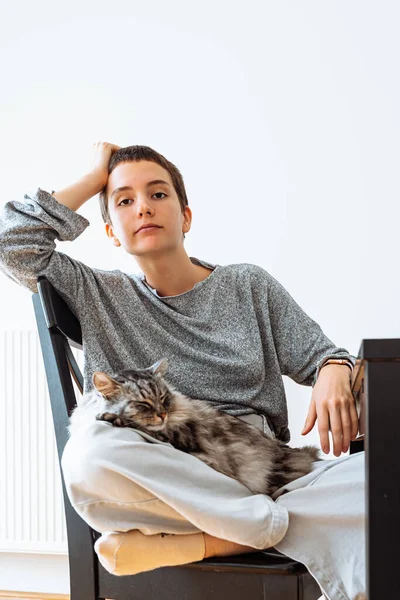 Portrait of teenager girl with short haircut, in casual clothes in gray tones, sitting with legs tucked under on chair at home, with homely fluffy gray sleeping cat in arms. Love and care for pets, teenager and cat friends spend time together