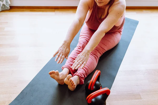 Beautiful middle-aged woman, plus size model, in sports top, sitting on mat at home and doing yoga, doing stretching, stretching arms to toes, making fold. View from above. Curvy woman takes care of body, does stretching, maintains healthy lifestyle