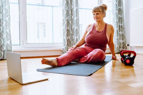 middle-aged woman with curvaceous, plump, blonde, sits on sports mat, does yoga, sports, at home oline. Active lifestyle, keeping body in good shape for middle-aged women