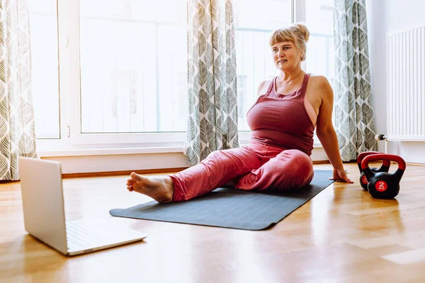 middle-aged woman with curvaceous, plump, blonde, sits on sports mat, does yoga, sports, at home oline. Active lifestyle, keeping body in good shape for middle-aged women
