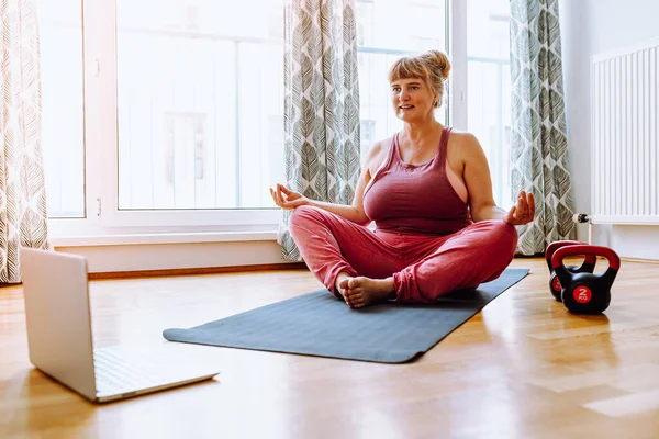 middle-aged woman, plus size, with blond hair tied in ponytail, sits on gymnastic mat in  lotus position with positive emotions, smiles, meditates, online relaxation training. Relaxation exercise in lotus position