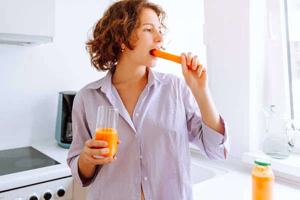 teenager girl eats fresh carrots with pleasure and holds carrot juice in glass in hand. Healthy proper nutrition, dietary breakfast, vitamins to improve vision. Happy cute girl eating carrot, vitamin A for good vision, health