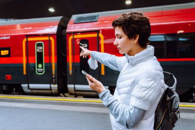 Kalın ceketli genç kız, istasyon peronuna gelen treni işaret ediyor, cep telefonunu elinde tutuyor. Online bilet rezervasyonu, tek kişilik tren yolculuğu.