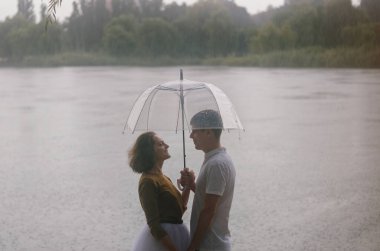 Kharkiv, Ukrayna. 09.19.2021. Şiddetli yağmurda şemsiyeyle nehrin kenarında yürüyen komik bir çiftin aşk hikayesi. Beyaz etekli, sarı kazaklı hoş bir adam ve kadın. Aile tatili ve birliktelik, randevu
