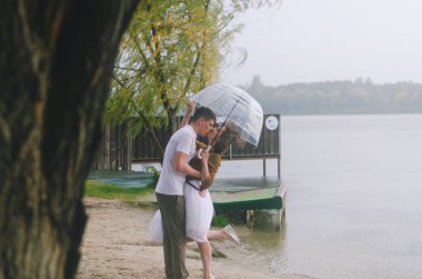 Kharkiv, Ukrayna. 09.19.2021. Şiddetli yağmurda şemsiyeyle nehrin kenarında yürüyen komik bir çiftin aşk hikayesi. Beyaz etekli, sarı kazaklı hoş bir adam ve kadın. Aile tatili ve birliktelik, randevu