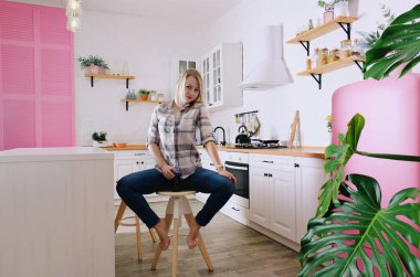 Genç, sarışın, 30 'lu yaşlarda, kot pantolonlu ve temiz mutfağında oturan ve rahatlatıcı gömlekli bir kadın. Sağlıklı ve mutlu bir yaşam tarzı. Boş zaman.