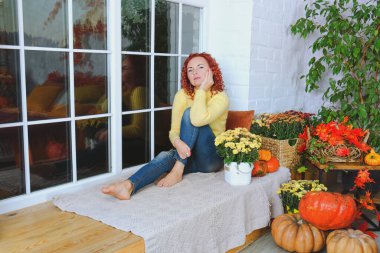 Güzel kızıl saçlı, kıvırcık saçlı, sıcak kazaklı bir kadın rahat bir evde yatakta oturuyor, gülüyor, eğleniyor. Sonbahar süslemeleri, portakal yaprakları, çiçekler, balkabakları olan ev.. 