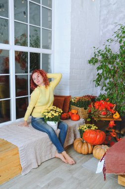 Güzel kızıl saçlı, kıvırcık saçlı, sıcak kazaklı bir kadın rahat bir evde yatakta oturuyor, gülüyor, eğleniyor. Sonbahar süslemeleri, portakal yaprakları, çiçekler, balkabakları olan ev.. 