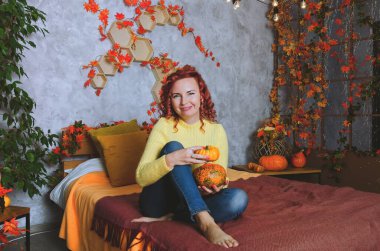 Güzel kızıl saçlı, kıvırcık saçlı, sıcak kazaklı bir kadın rahat bir evde yatakta oturuyor, gülüyor, eğleniyor. Sonbahar süslemeleri, portakal yaprakları, çiçekler, balkabakları olan ev.. 