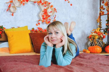 Sarı süveterli, genç, sarışın bir kadın, sonbahar süsleri, turuncu balkabakları, sonbahar çiçekleri olan rahat evinde, pencere kenarındaki koltuğa oturur. Sonbahar sezonu. Çay fincanı. 