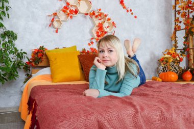 Sarı süveterli, genç, sarışın bir kadın, sonbahar süsleri, turuncu balkabakları, sonbahar çiçekleri olan rahat evinde, pencere kenarındaki koltuğa oturur. Sonbahar sezonu. Çay fincanı. 